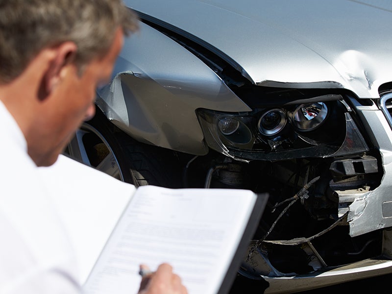 A man examines a car as he writes on a clipboard in front of it.