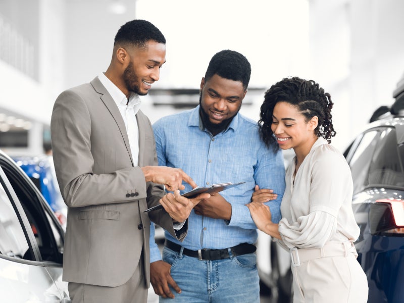A group of three individuals reviewing a tablet in a car dealership setting.